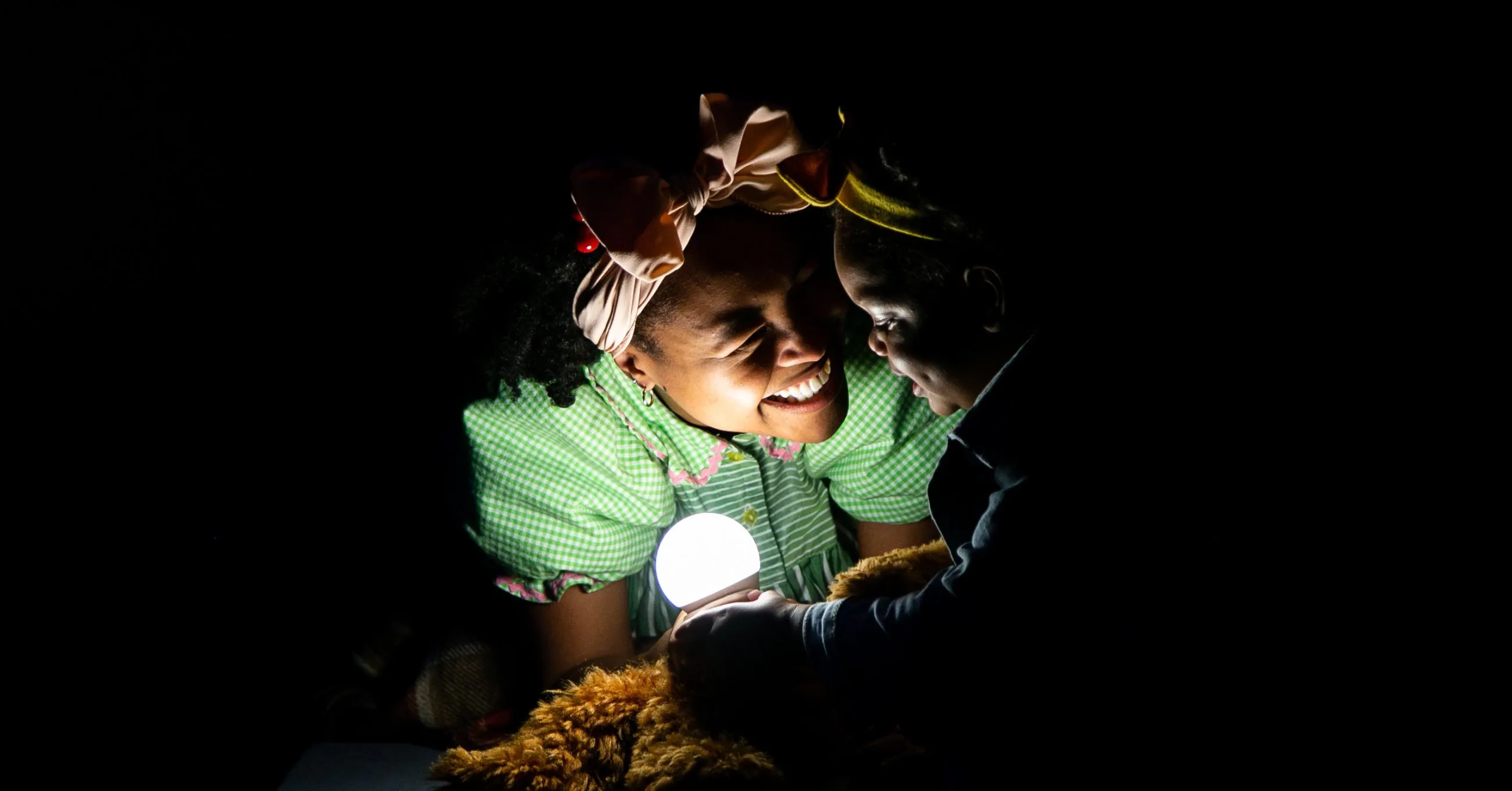 Two children wearing costumes smile at each other while holding a glowing light in the dark, with their faces illuminated.