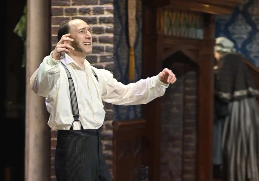 A man in vintage clothing gestures dramatically on stage, wearing a white shirt and suspenders, with another person blurred in the background near a brick wall and wooden cabinet.