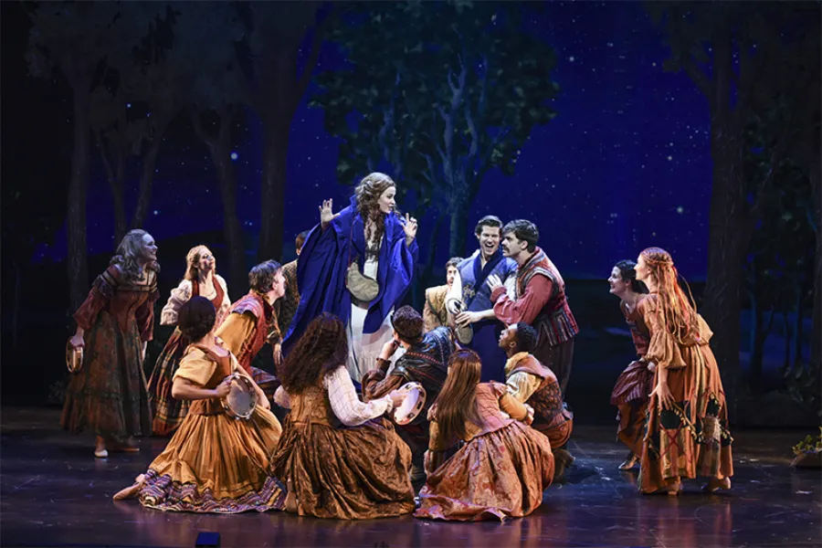 A group of actors in colorful costumes perform a lively scene on stage, with one central figure in blue surrounded by others, set against a backdrop of trees and a starry night sky.