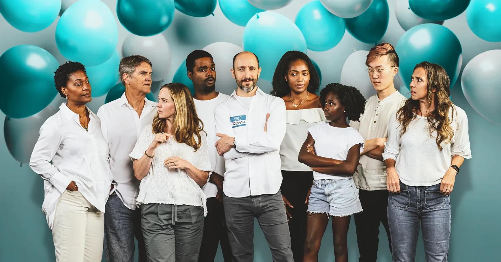 A diverse group of nine people in casual white clothing stands in front of a teal balloon backdrop, looking in various directions.