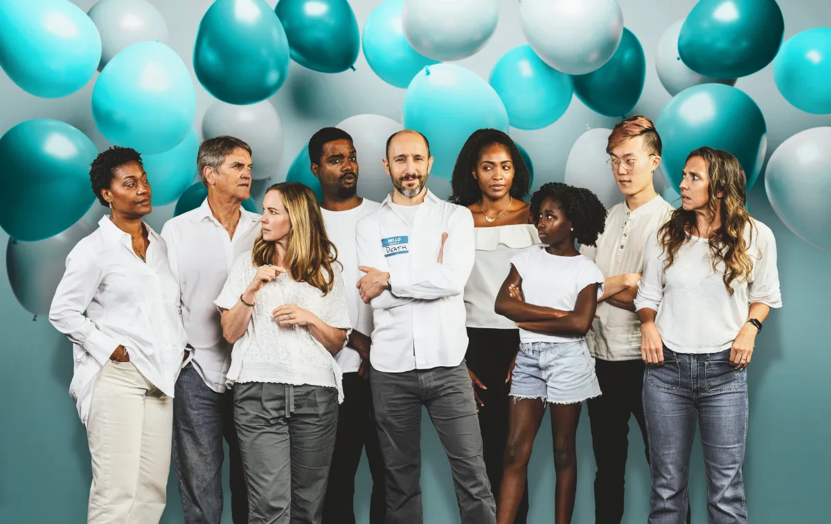 A diverse group of people wearing white clothes stands in front of a wall decorated with blue and white balloons.