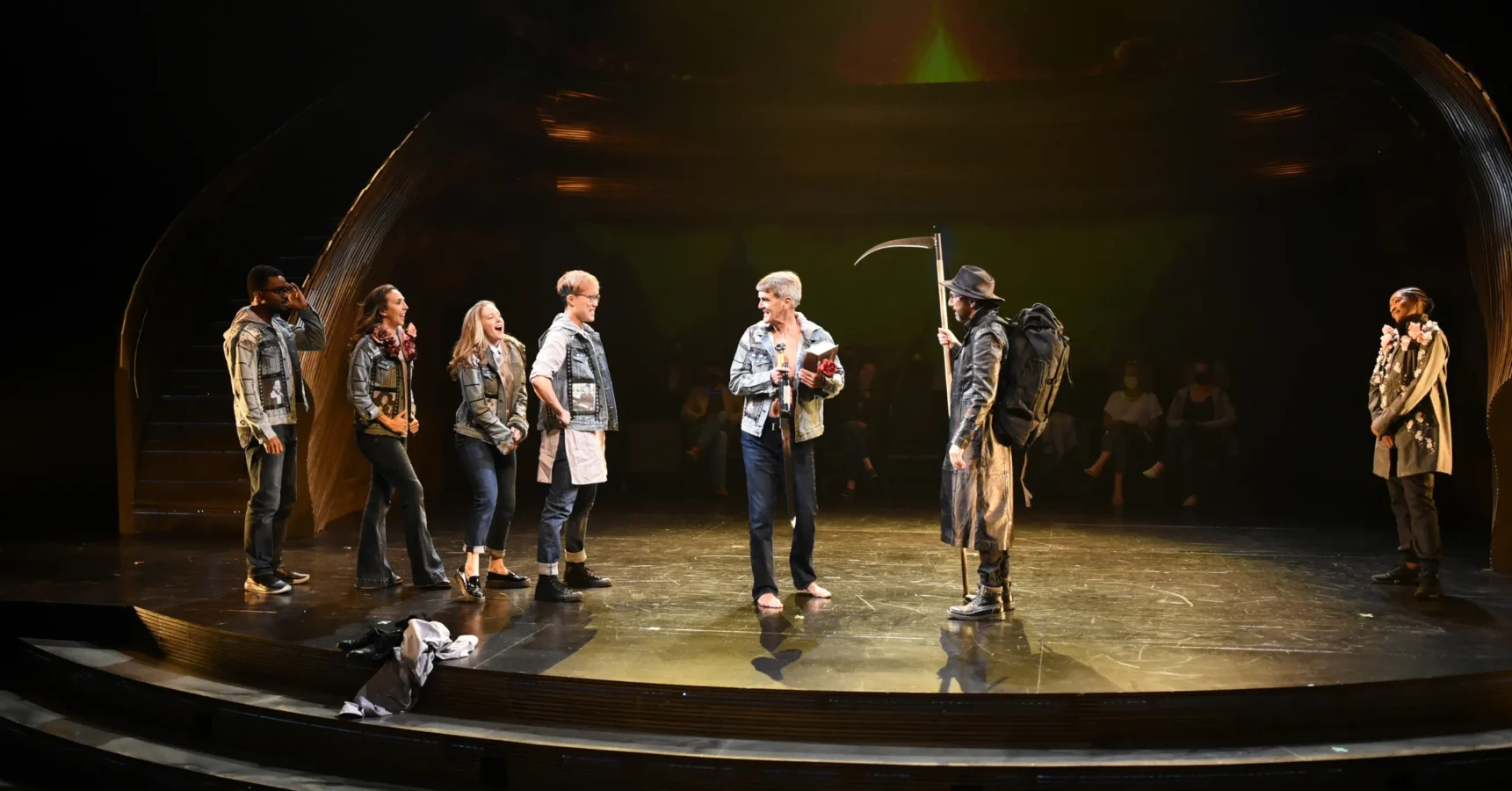 A group of actors stand on a stage; one holds a scythe and wears dark clothing, while the others face him, dressed in casual outfits and jackets.
