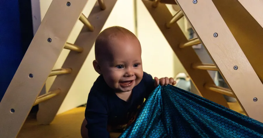 A baby in dark clothing crawls under a wooden climbing structure, smiling and pulling on a blue patterned fabric.
