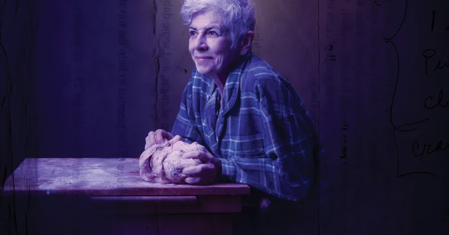 Older woman in a plaid shirt sits at a wooden table, shaping a braided loaf of dough, with a soft purple light illuminating the scene.