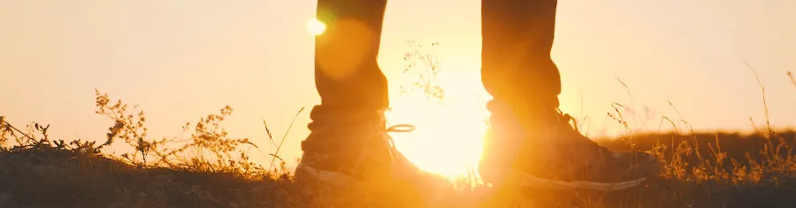 Close-up of a person's shoes standing on grass during sunset, with bright sunlight visible in the background.