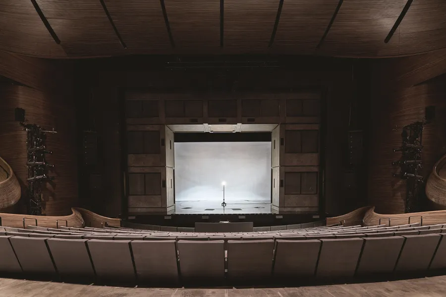 An empty theater with tiered seating faces a stage lit by a single spotlight centered on a microphone stand, with wooden walls and ceiling visible.