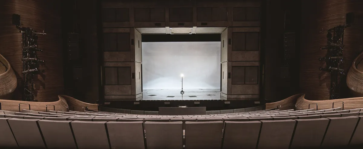 Empty theater auditorium with rows of seats facing a stage, featuring a single lit microphone stand at the center under white lighting.