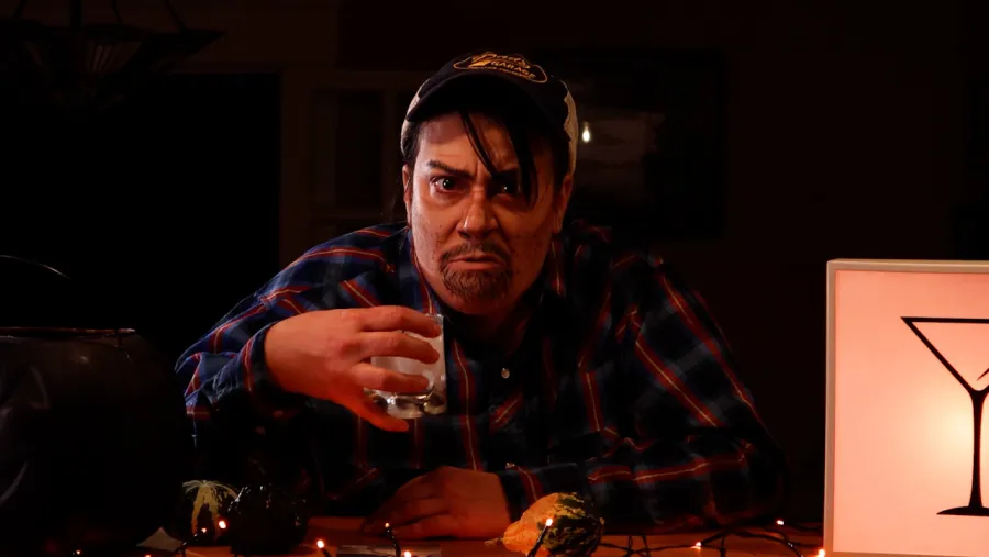 Person in a plaid shirt and baseball cap holds a glass of clear liquid, sitting at a table with decorative gourds and orange string lights, next to a sign with a martini glass symbol.