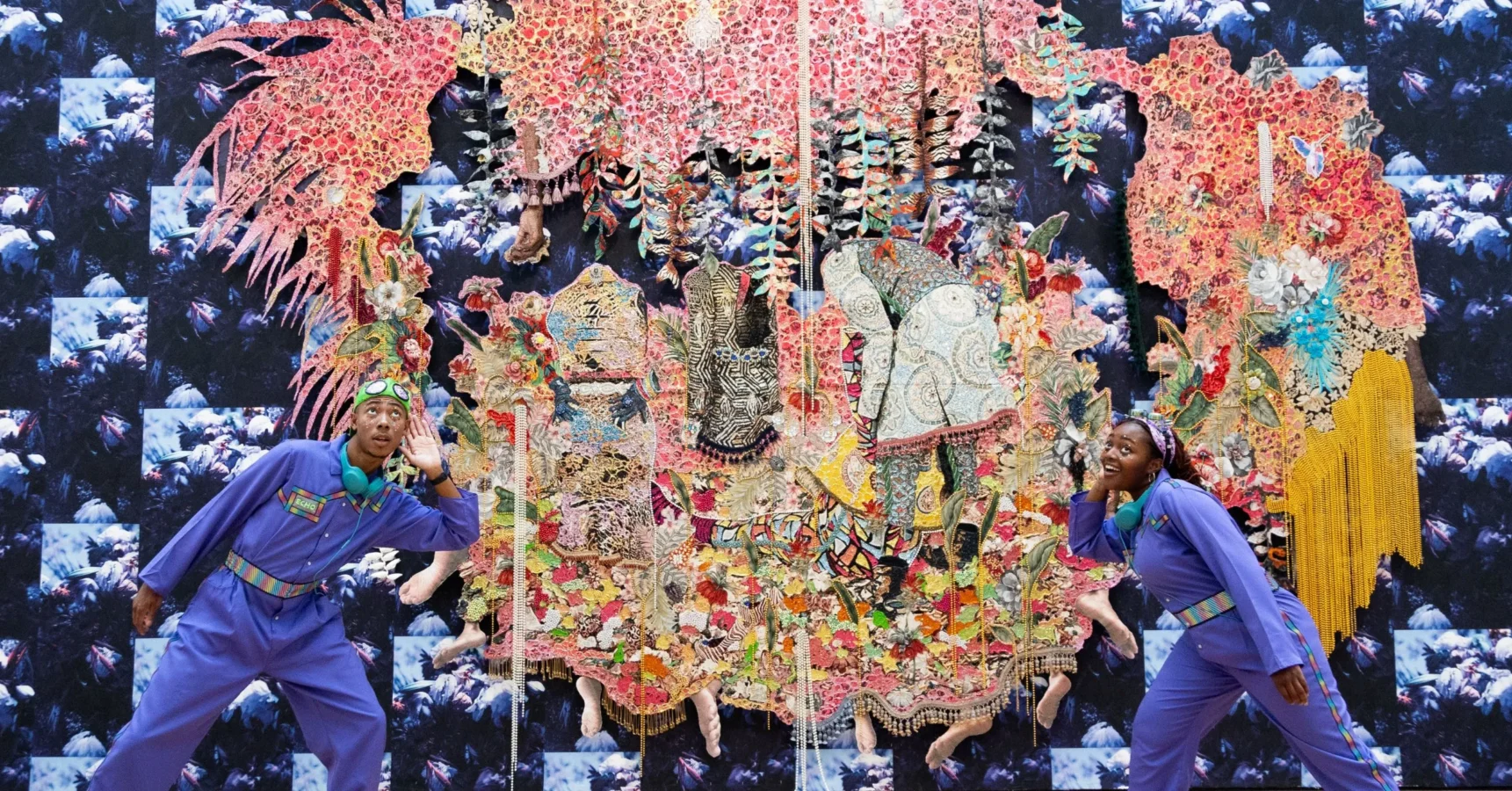 Two people in matching blue outfits pose playfully in front of a large, colorful, intricate textile art piece with a patterned backdrop.