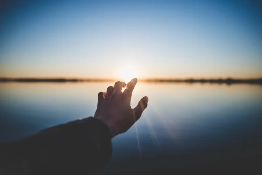 A hand reaching toward the setting sun over a calm, reflective body of water under a clear sky.