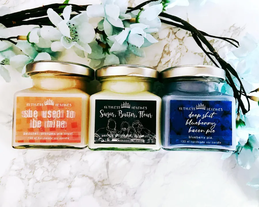 Three scented candles in glass jars with silver lids, each labeled with a different scent and pie-themed name, placed on a white marble surface with white flowers nearby.