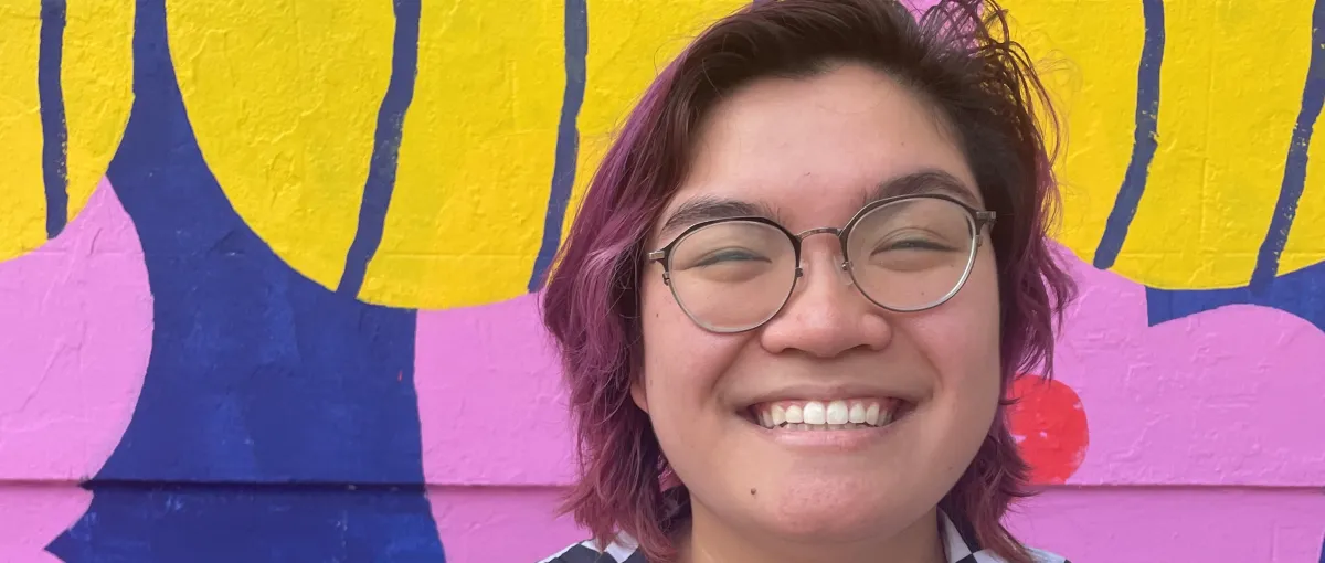 Person with glasses and short, reddish hair smiling in front of a colorful, abstract mural with yellow, blue, and pink shapes.