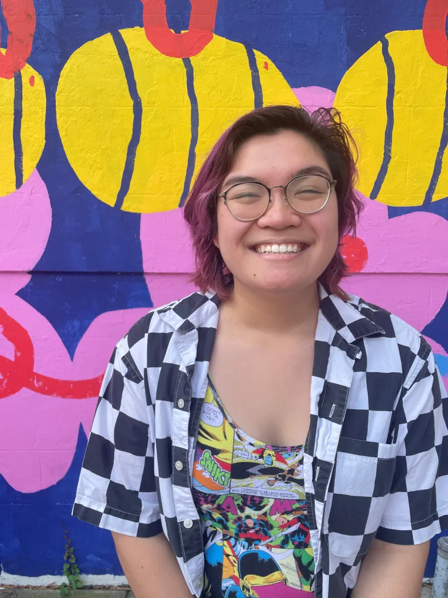 Person with short hair and glasses smiling in front of a colorful mural with yellow, pink, and blue shapes. They are wearing a black-and-white checkered shirt and a patterned top.