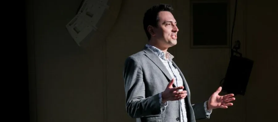 A man in a grey blazer and white shirt stands on stage, gesturing with his hands as he speaks, with stage lighting focused on him.