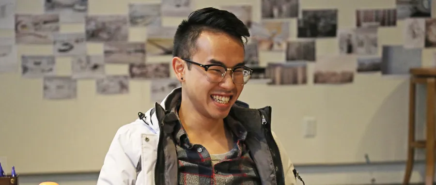A person wearing glasses and a plaid shirt smiles while sitting indoors; blurred photos and sketches are displayed on the wall behind them.