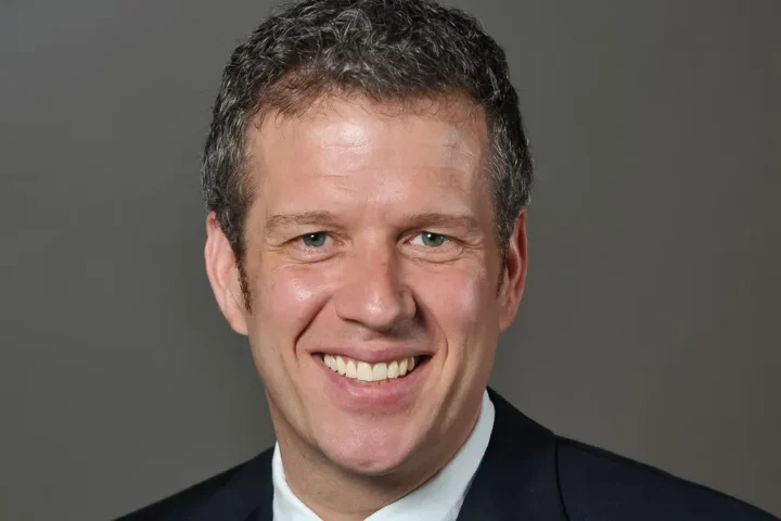 A man in a dark suit and white shirt smiles at the camera against a plain, gray background.