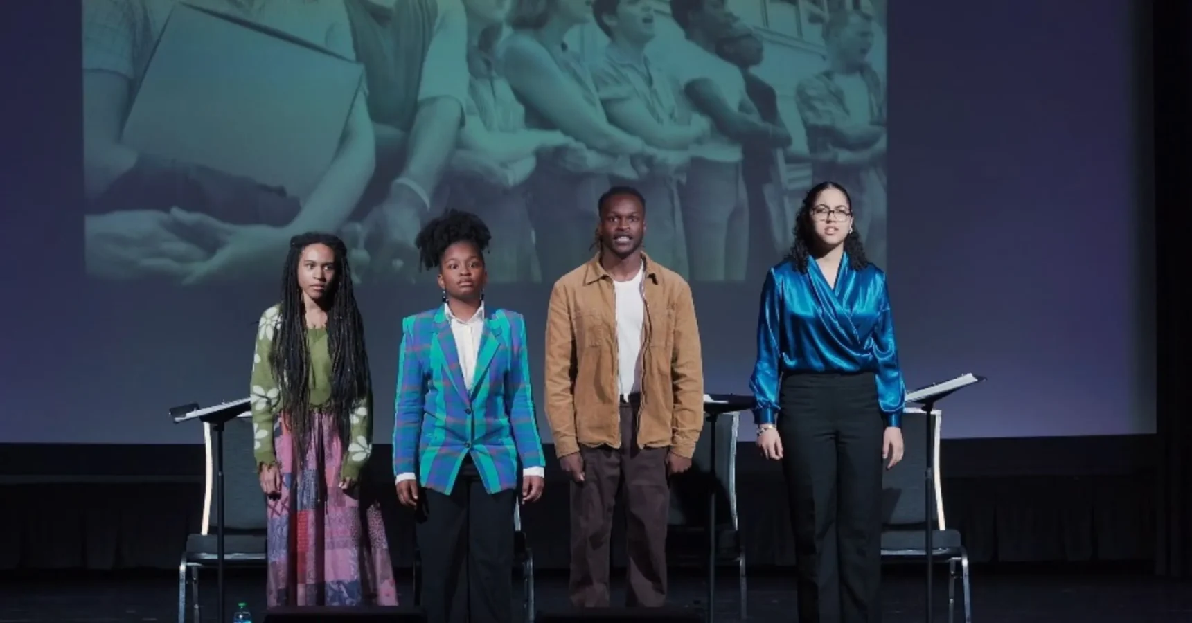 Four people stand side by side on stage in front of chairs and music stands, with a black-and-white historical photo of people holding hands projected behind them.