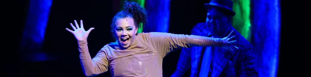A performer with curly hair and expressive makeup poses with raised hands on stage, illuminated by blue and green lights, while another person stands in the background.