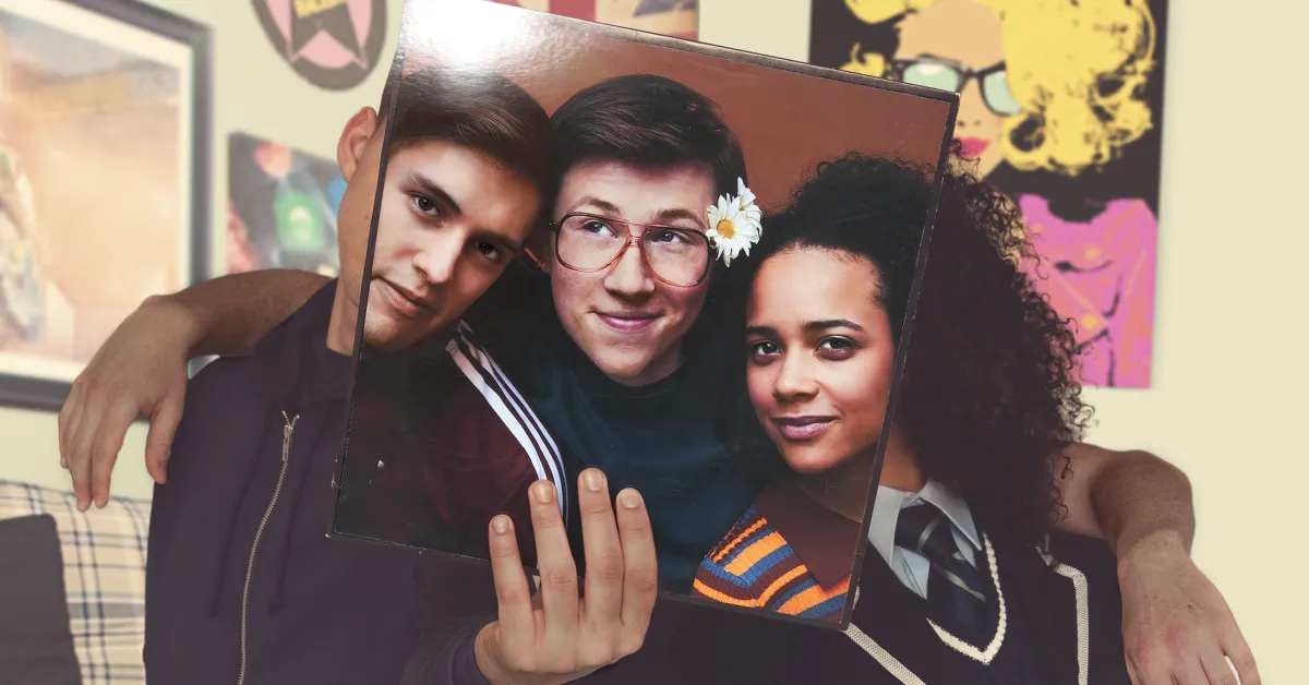 Three people sit closely together, holding a photo of three different people in front of their faces, aligning the faces with their own bodies in a room decorated with pop art posters.