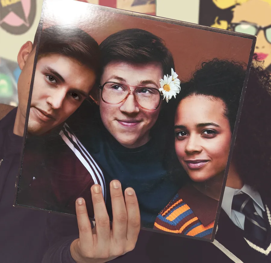 A person holds a vinyl album cover in front of their face, aligning the cover's faces with their own, creating an illusion where the cover and real people blend together.