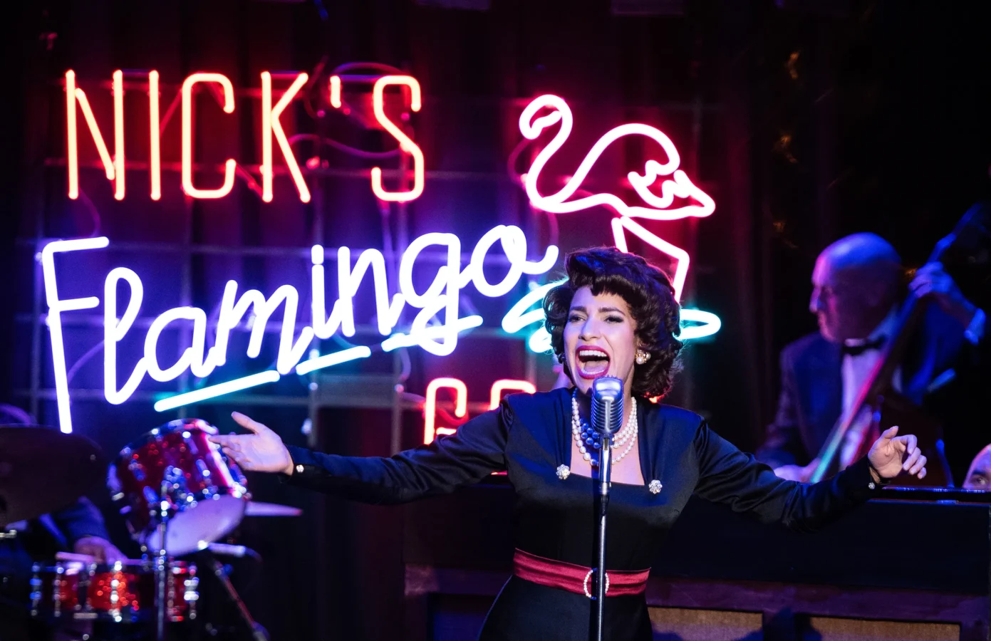 A woman in vintage attire sings into a retro microphone on stage, with a neon sign reading "Nick's Flamingo" and musicians playing in the background.
