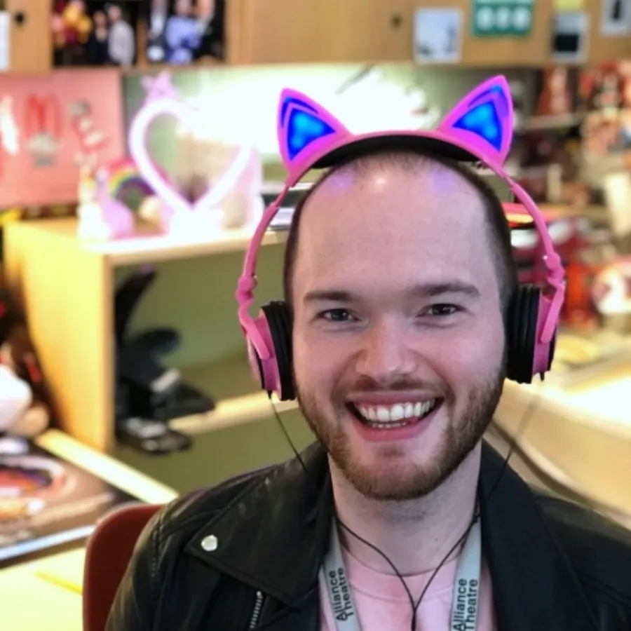 A person wearing light-up cat ear headphones and a lanyard smiles at the camera in a brightly decorated office space.