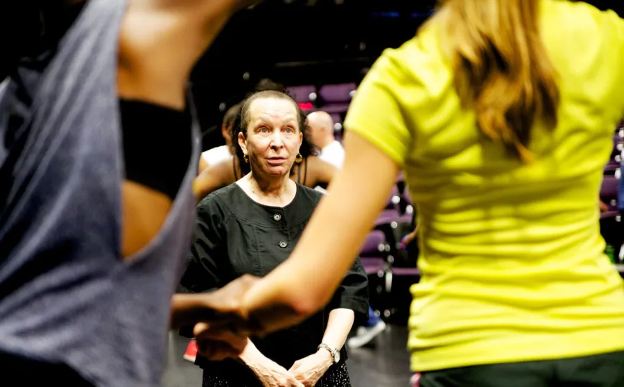 A woman in a black shirt stands indoors, observing two people holding hands in the foreground.