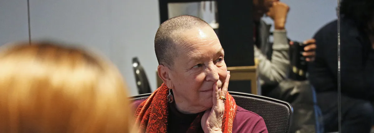 An older woman with a shaved head and wearing a red scarf rests her chin on her hand, looking to the side. Other people are visible in the blurred background.