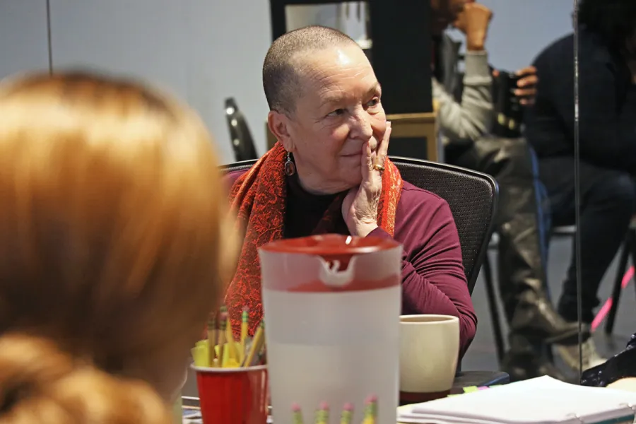 A person with a shaved head and red scarf sits at a table with a pitcher, pencils, and a mug, resting their hand on their cheek and looking to the side.