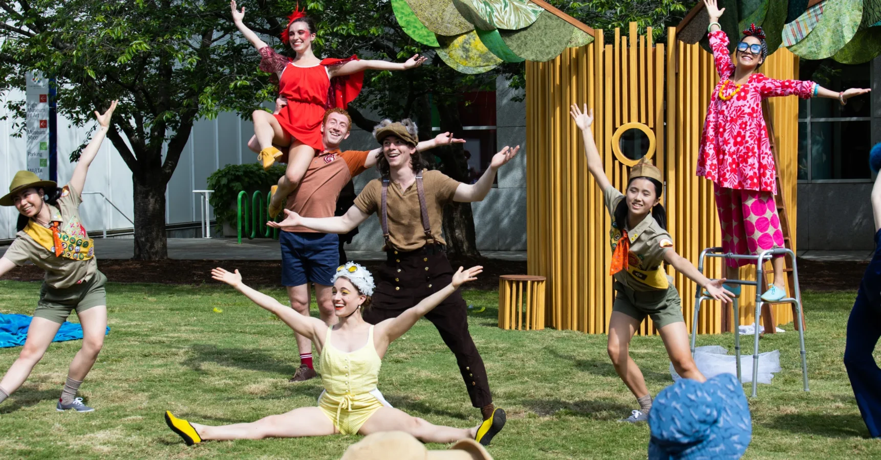 Performers in colorful costumes pose in a cheerful outdoor scene, with some standing and others mid-dance or doing splits on the grass.