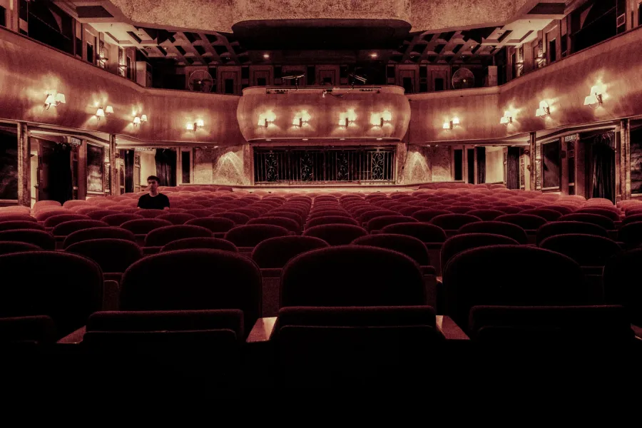 Rows of empty red theater seats in a dimly lit auditorium, with a single person sitting alone near the left side of the audience.