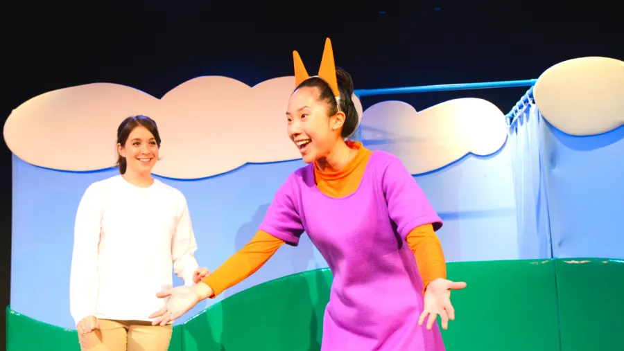 Two actors perform on stage; one wears a purple dress and orange headpiece, while the other stands in white clothes, with a cartoonish outdoor backdrop behind them.