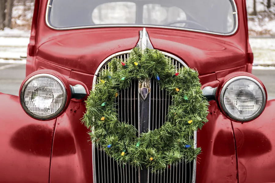 A close-up of a vintage red car with a green Christmas wreath decorated with lights attached to the front grille.