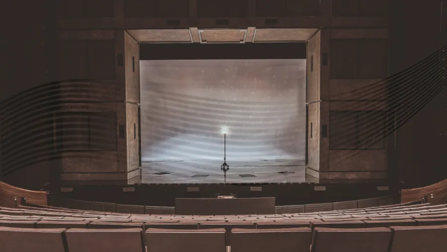 A theater stage with a closed curtain, empty seats in the foreground, and a single standing microphone at the center of the stage.