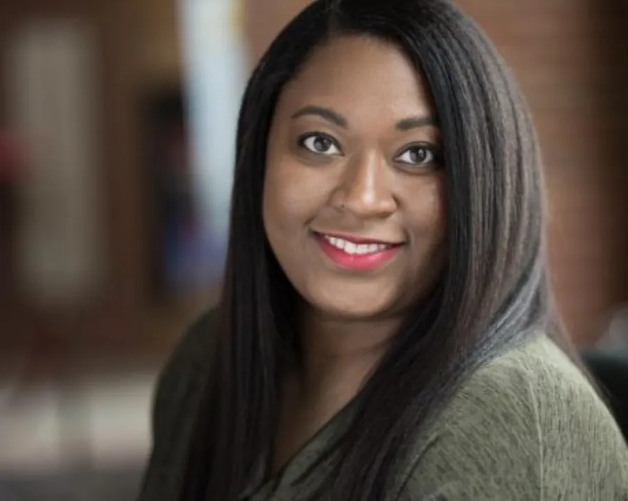 A woman with long straight hair and a green top smiles at the camera in an indoor setting with a blurred background.