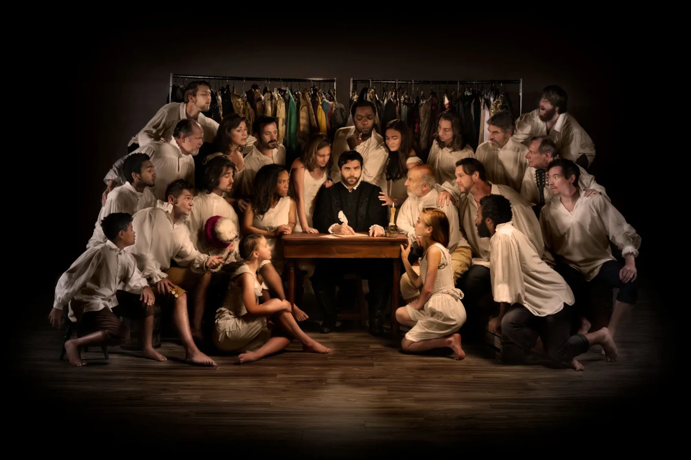 A group of people dressed in white shirts gathers around a man seated at a table, with clothing racks in the background and dramatic lighting highlighting the scene.