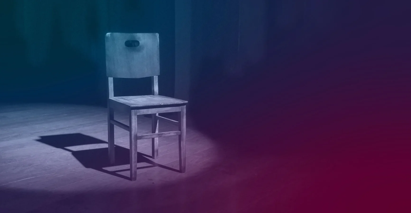 A single wooden chair sits on a wooden floor in a spotlight, casting a shadow. The background is dark with blue and purple tones.