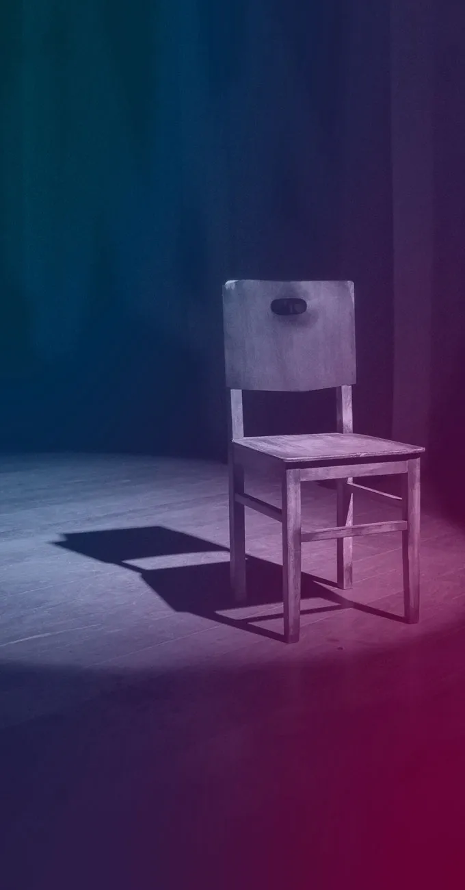 A single wooden chair with a rectangular cutout in the backrest is positioned on a dimly lit stage, casting a shadow on the floor.