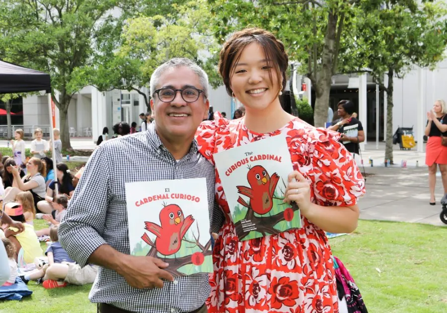 Two people stand outdoors holding children's books with a red cardinal on the cover; a seated crowd is visible in the background.