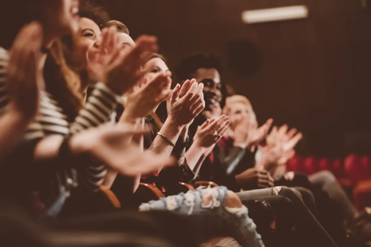 A group of people sitting in theater seats clapping and facing the stage, with some smiling and appearing engaged with the performance.