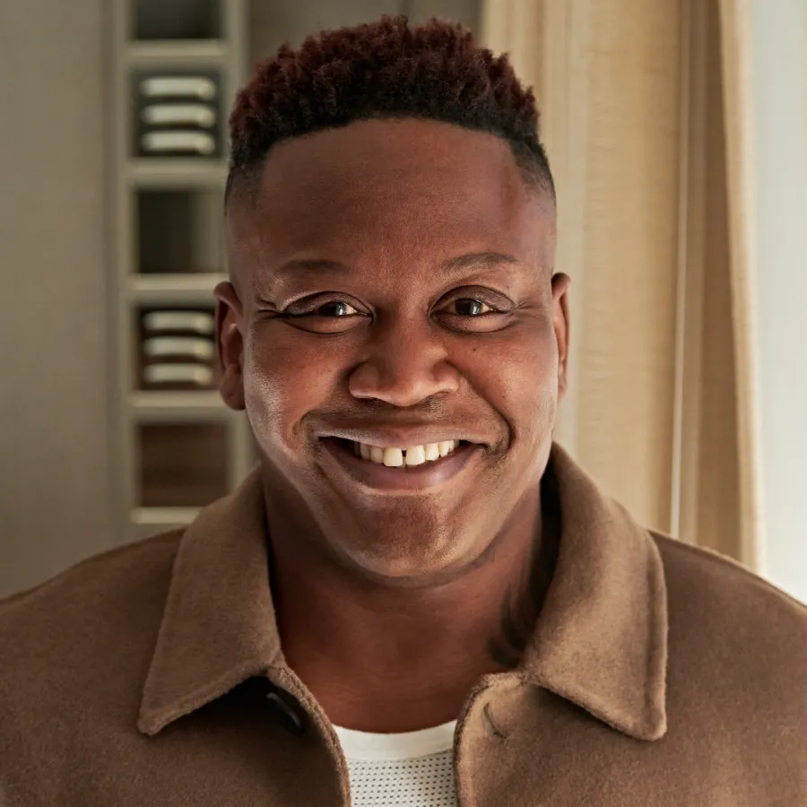 A man with short, curly hair and a brown jacket smiles while standing indoors in front of shelves and a curtain.