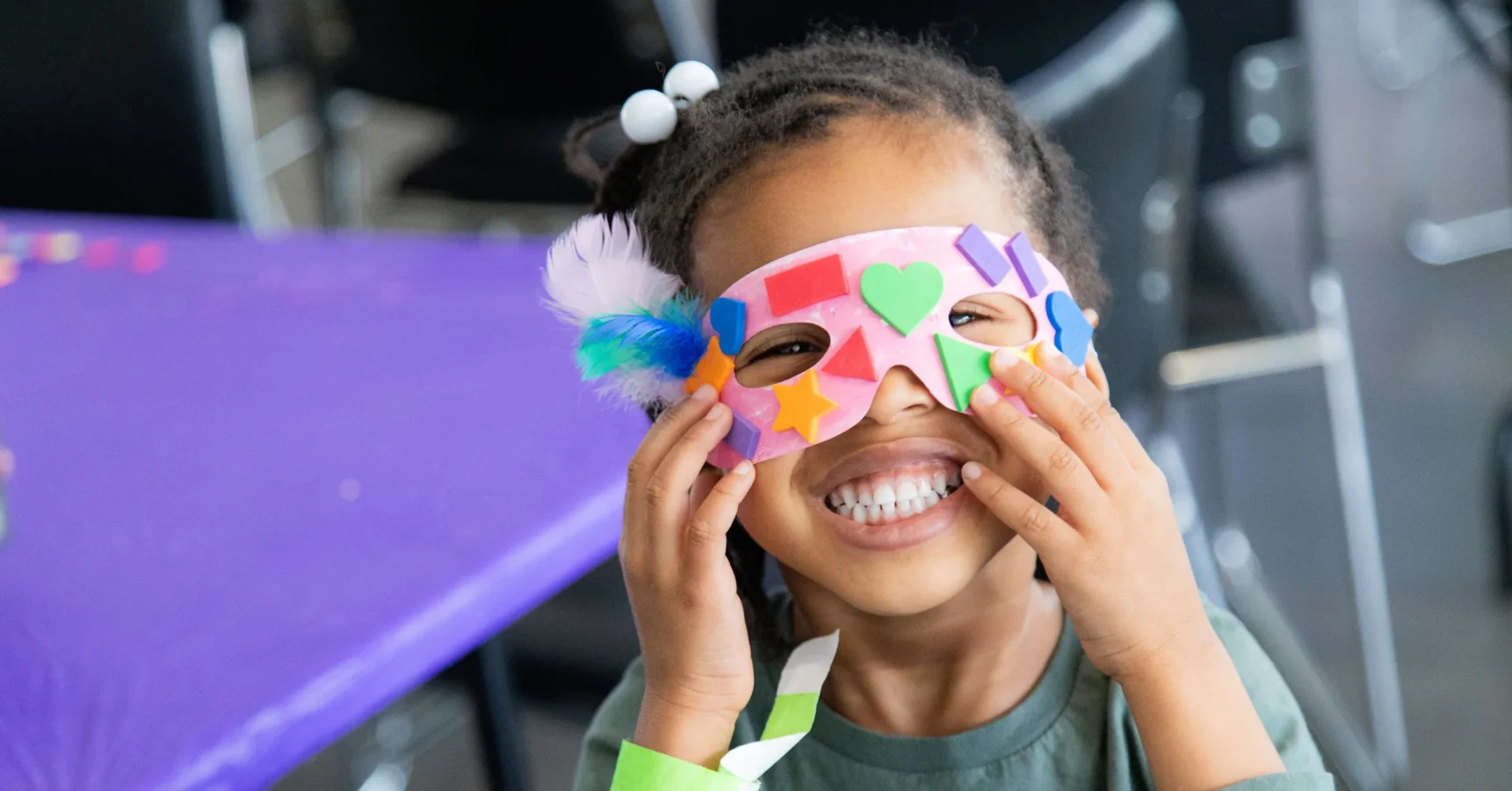 Smiling child holding a colorful, handmade paper mask decorated with foam shapes and feathers, seated at a table with a purple tablecloth.