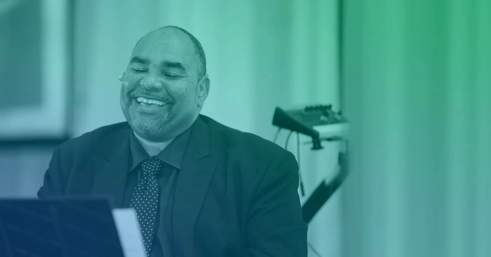 A man in a suit and tie smiles while sitting at a keyboard or piano, with electronic music equipment visible in the background.