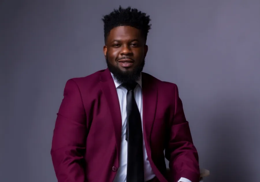 A man with short curly hair and a beard wearing a maroon suit, white shirt, and black tie sits against a plain gray background.