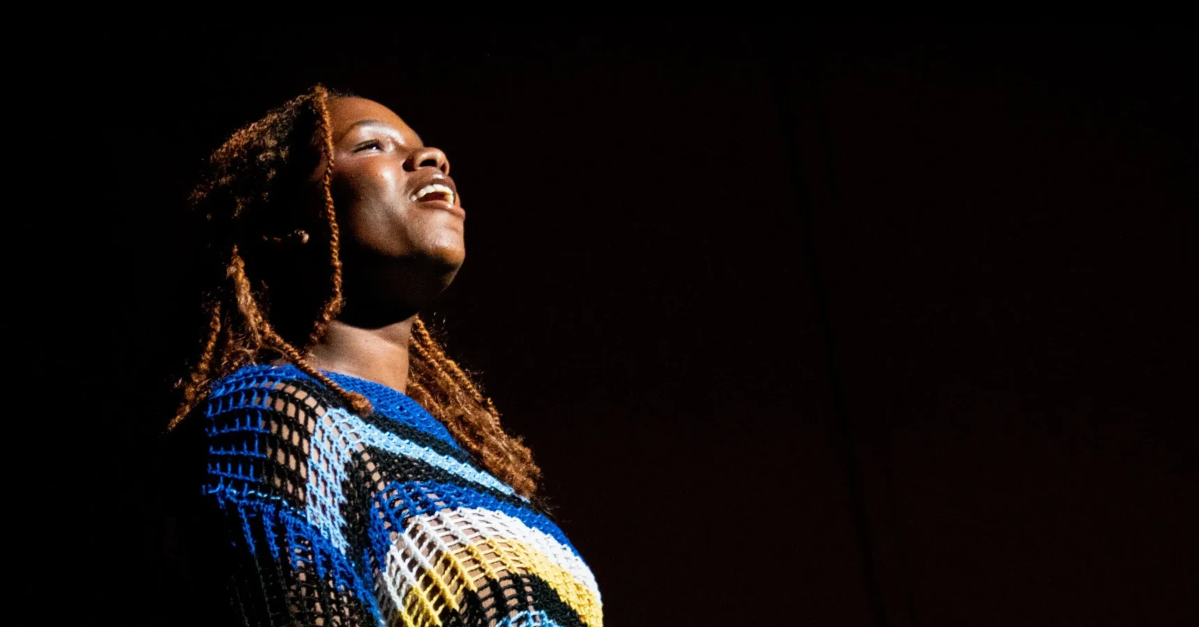 A person with long hair sings or speaks on stage under a spotlight, wearing a blue, yellow, and white patterned top against a dark background.
