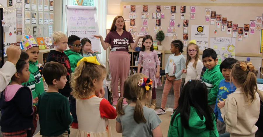 A teacher stands in a circle with young children in a classroom, leading an activity; educational posters and student artwork decorate the walls.