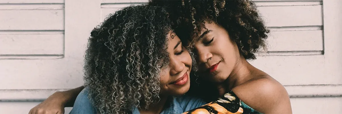 Two women with curly hair sit closely together, eyes closed and heads touching, appearing relaxed and content in front of a white wooden wall.