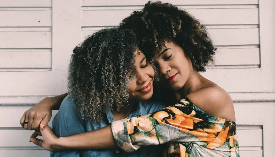 Two women with curly hair embrace in front of a white wooden wall, both smiling with eyes closed in a relaxed, affectionate pose.