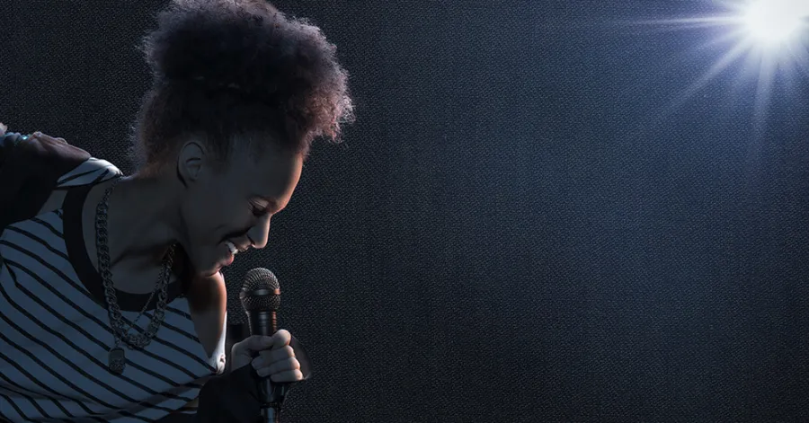 A person with curly hair holds a microphone and sings on stage under a bright spotlight, with a dark background.