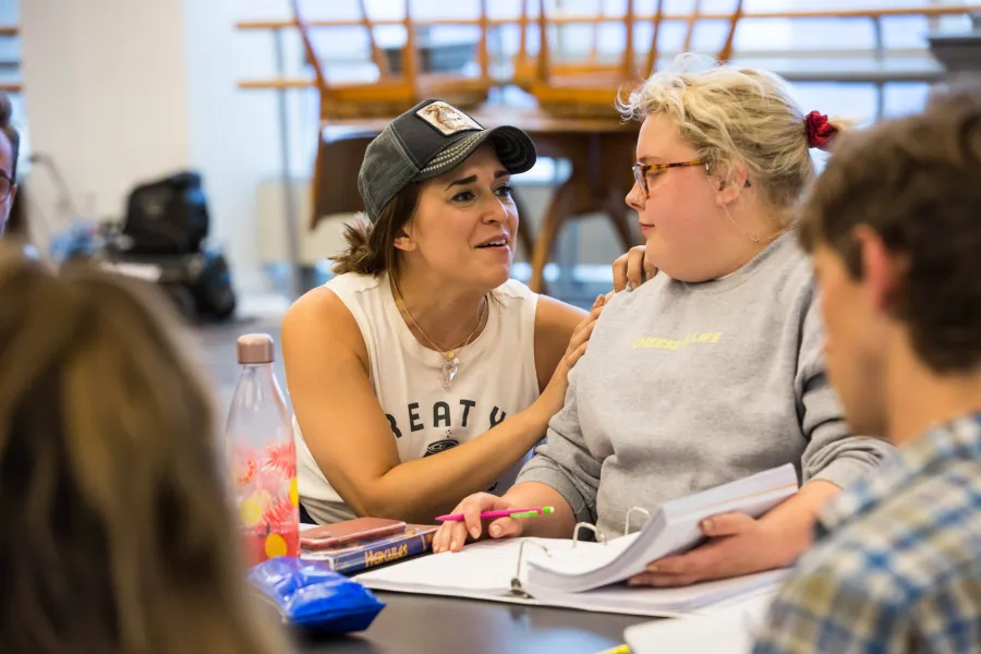 Two women sit at a table with notebooks and pens; one leans in to talk to the other, who is listening, in a classroom or study setting.